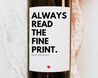 May include: A wine bottle with a white label that says "ALWAYS READ THE FINE PRINT. WE'RE PREGNANT!" with a red heart at the bottom.