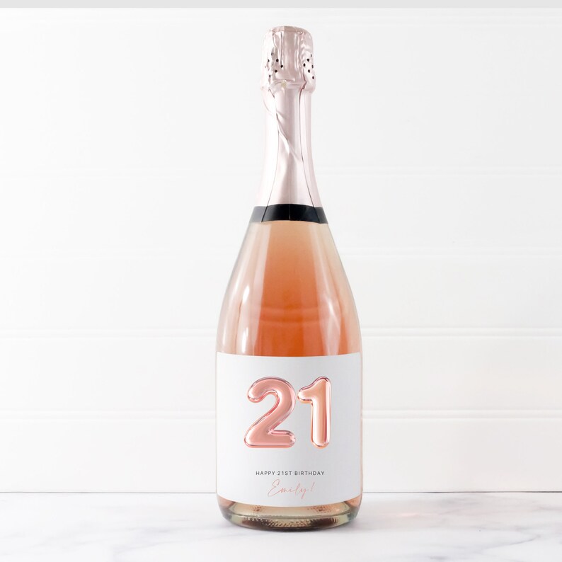 May include: A bottle of pink champagne with a white label that says "Happy 21st Birthday Emily!" and features a rose gold number 21.