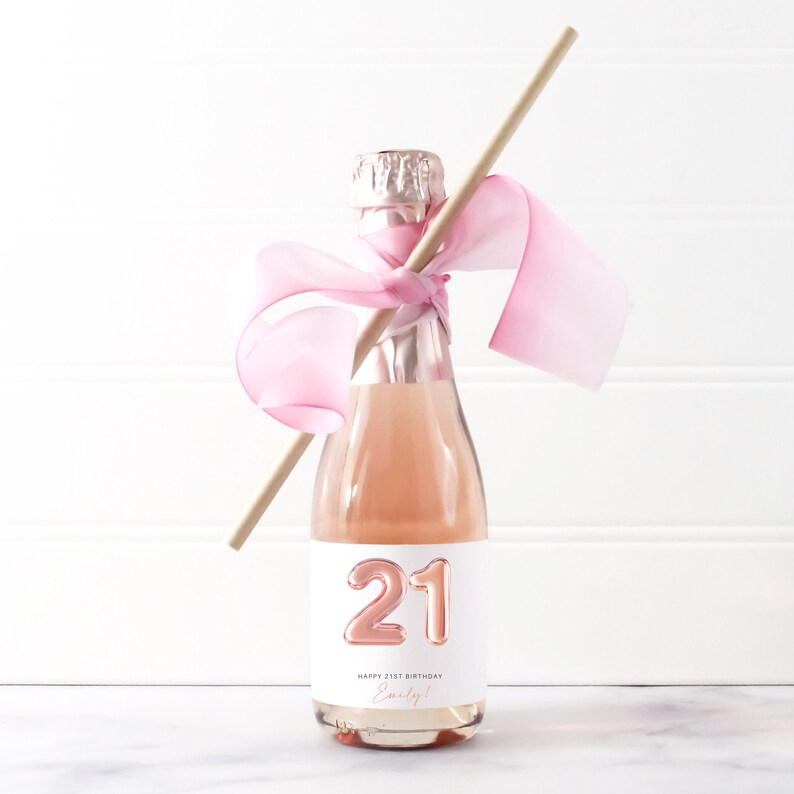 May include: A small bottle of pink champagne with a white label that says "Happy 21st Birthday Emily!" and has a large, rose gold number "21" on it. The bottle is tied with a pink ribbon and a wooden straw.