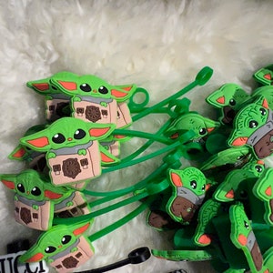 Op de afbeelding: Een groep groene siliconen kabelorganizers met een cartoonfiguur dat een doos vasthoudt. Het figuur heeft grote ogen, een kleine mond en twee oren. De organizers zijn bevestigd aan groene koorden.