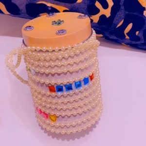 Op de afbeelding: Een kleine, ronde, perzikkleurige container met een kralen deksel en een decoratieve band van kleurrijke juwelen. De container is omgeven door een string van witte parels.