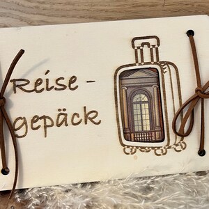 May include: A wooden travel journal with a brown leather strap and the text "Reise - Gep&auml;ck" on the front. The journal has a cutout of a suitcase with a window.