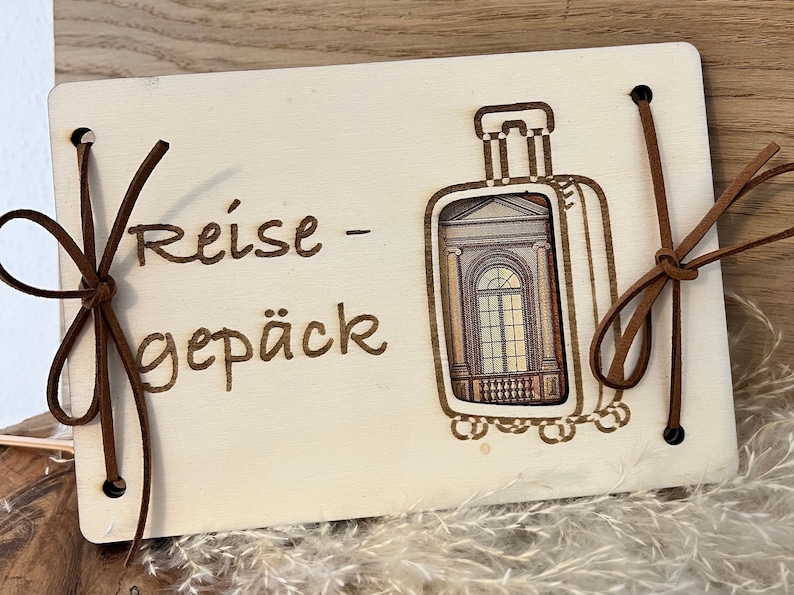 May include: A wooden travel journal with a brown leather strap and the text "Reise - Gep&auml;ck" on the front. The journal has a cutout of a suitcase with a window.