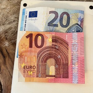 May include: A white card with two Euro banknotes: a 20 Euro note and a 10 Euro note. The 20 Euro note is partially visible, while the 10 Euro note is fully visible. The background is a textured, light brown surface.