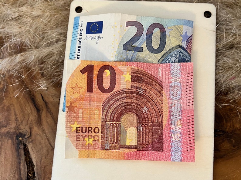 May include: A white card with two Euro banknotes: a 20 Euro note and a 10 Euro note. The 20 Euro note is partially visible, while the 10 Euro note is fully visible. The background is a textured, light brown surface.