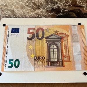 May include: A 50 euro bill with a building and stars on it. The bill is framed in a white frame with holes in the corners.