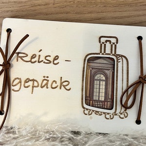 May include: A wooden travel journal with a brown leather strap and the text "Reise - Gep&auml;ck" engraved on the front. The journal has a cutout of a suitcase with a window view.