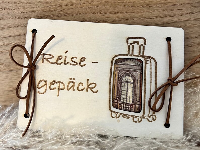 May include: A wooden travel journal with a brown leather strap and the text "Reise - Gep&auml;ck" engraved on the front. The journal has a cutout of a suitcase with a window view.