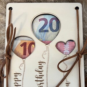 May include: Wooden birthday card with cut-out balloon shapes holding currency. The card has brown leather ties and the words "Happy Birthday" are etched on the front. The balloons are filled with the numbers 10 and 20.