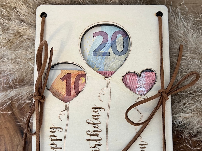 May include: Wooden birthday card with cut-out balloon shapes holding currency. The card has brown leather ties and the words "Happy Birthday" are etched on the front. The balloons are filled with the numbers 10 and 20.