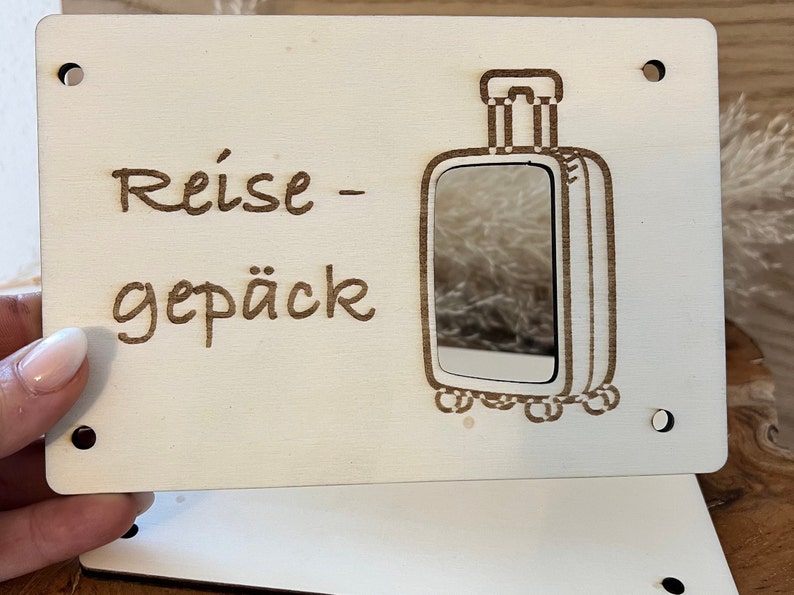 May include: A white wooden sign with the text "Reise-gep&auml;ck" and a cutout of a suitcase with a mirror inside.