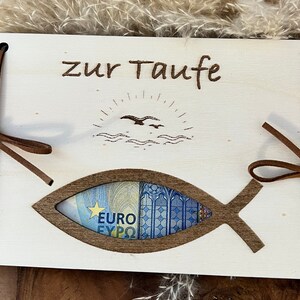 Op de afbeelding: Een houten doopcadeaukaarthouder met een visvormige uitsparing. De woorden "zur Taufe" zijn bovenaan ge&euml;tst, met een zon- en vogelontwerp. Bruine leren banden bevestigen de kaarthouder.
