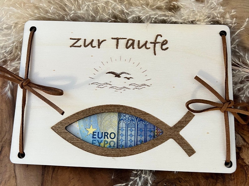 Op de afbeelding: Een houten doopcadeaukaarthouder met een visvormige uitsparing. De woorden "zur Taufe" zijn bovenaan ge&euml;tst, met een zon- en vogelontwerp. Bruine leren banden bevestigen de kaarthouder.