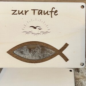 Op de afbeelding: Lichtgekleurde houten plaat met de woorden "zur Taufe" gegraveerd bovenaan. Onder de tekst staat een zon- en vogelontwerp en een visvormige uitsparing. De plaat heeft vier kleine gaten in de hoeken.