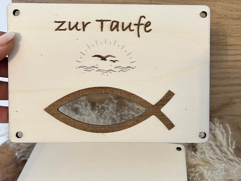 Op de afbeelding: Lichtgekleurde houten plaat met de woorden "zur Taufe" gegraveerd bovenaan. Onder de tekst staat een zon- en vogelontwerp en een visvormige uitsparing. De plaat heeft vier kleine gaten in de hoeken.
