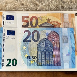 Op de afbeelding: Een witte rechthoekige houder met twee eurobiljetten: een biljet van 50 euro en een biljet van 20 euro. Het biljet van 20 euro is blauw en heeft een gotisch vensterontwerp. Het biljet van 50 euro is oranje.