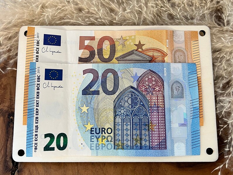 Op de afbeelding: Een witte rechthoekige houder met twee eurobiljetten: een biljet van 50 euro en een biljet van 20 euro. Het biljet van 20 euro is blauw en heeft een gotisch vensterontwerp. Het biljet van 50 euro is oranje.