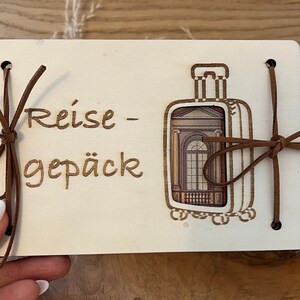 May include: A white wooden sign with the text "Reise - Gep&auml;ck" and a brown leather luggage tag with a window design. The sign has brown leather ties on the sides.