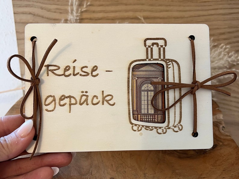 May include: A white wooden sign with the text "Reise - Gep&auml;ck" and a brown leather luggage tag with a window design. The sign has brown leather ties on the sides.