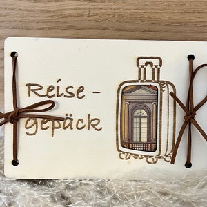 May include: A white wooden sign with the text "Reise - Gep&auml;ck" and a brown leather luggage tag with a picture of a building with a window. The sign has two brown leather straps tied around it.