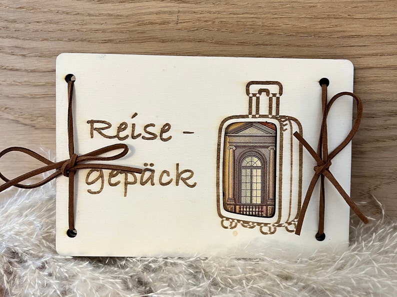 May include: A white wooden sign with the text "Reise - Gep&auml;ck" and a brown leather luggage tag with a picture of a building with a window. The sign has two brown leather straps tied around it.