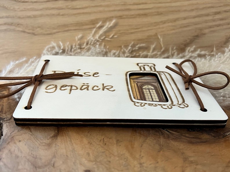 May include: White wooden box with a brown leather strap and the text "Reise-gep&auml;ck" on the front. The box has a cutout of a suitcase with a window.