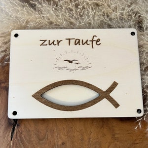 Op de afbeelding: Een lichtgekleurd houten bord met de Duitse tekst "zur Taufe" en een vissymbool. Het bord heeft ook een gegraveerde afbeelding van een zon, golven en vogels. Het bord heeft vier zwarte schroefgaten in elke hoek.