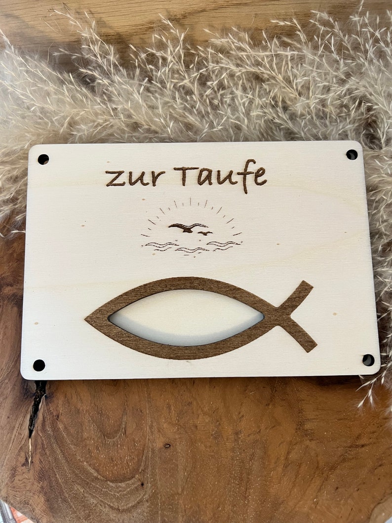 Op de afbeelding: Een lichtgekleurd houten bord met de Duitse tekst "zur Taufe" en een vissymbool. Het bord heeft ook een gegraveerde afbeelding van een zon, golven en vogels. Het bord heeft vier zwarte schroefgaten in elke hoek.