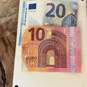May include: A wooden surface with two Euro banknotes: a 20 Euro note and a 10 Euro note. The 20 Euro note is blue, grey, and white, while the 10 Euro note is orange, yellow, and red. The notes are partially inserted into a light-coloured wooden holder.