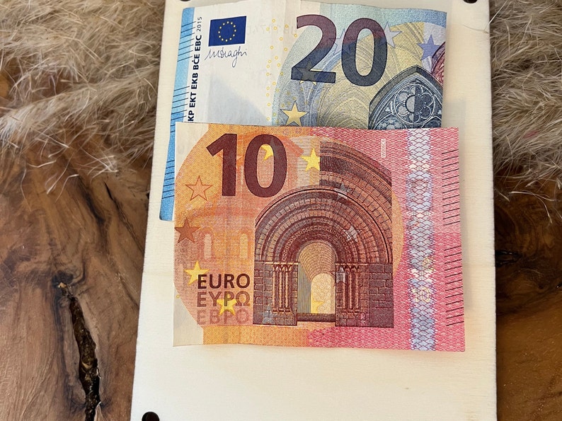 May include: A wooden surface with two Euro banknotes: a 20 Euro note and a 10 Euro note. The 20 Euro note is blue, grey, and white, while the 10 Euro note is orange, yellow, and red. The notes are partially inserted into a light-coloured wooden holder.