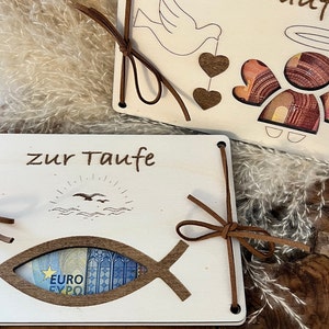Op de afbeelding: Twee houten doopcadeaus met de woorden "zur Taufe" gegraveerd. E&eacute;n heeft een visvormige uitsparing met een eurobiljet, de andere een engel- en duifontwerp. Beide zijn vastgebonden met bruine leren koorden.