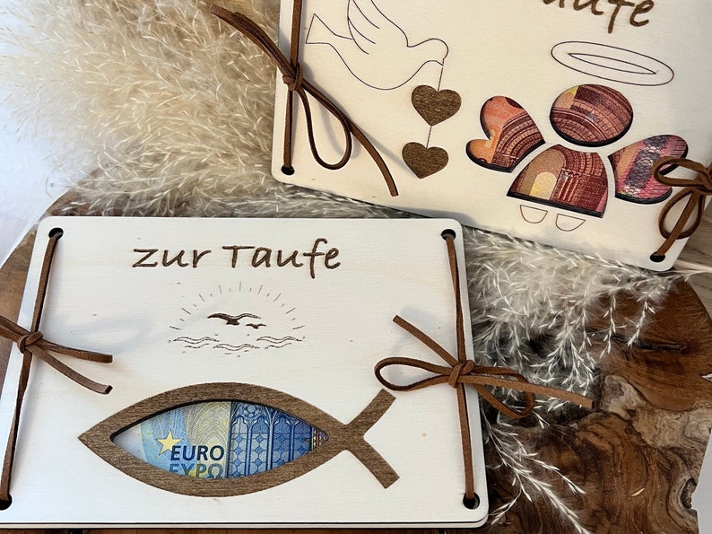 Op de afbeelding: Twee houten doopcadeaus met de woorden "zur Taufe" gegraveerd. E&eacute;n heeft een visvormige uitsparing met een eurobiljet, de andere een engel- en duifontwerp. Beide zijn vastgebonden met bruine leren koorden.