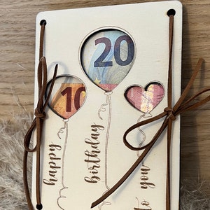 May include: A wooden birthday card with balloon cutouts displaying currency. The card has the words "happy birthday to you" and features brown leather ties. The balloons are in the shape of a heart and two balloons.