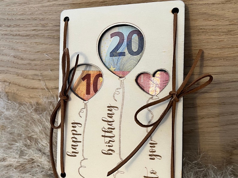 May include: A wooden birthday card with balloon cutouts displaying currency. The card has the words "happy birthday to you" and features brown leather ties. The balloons are in the shape of a heart and two balloons.