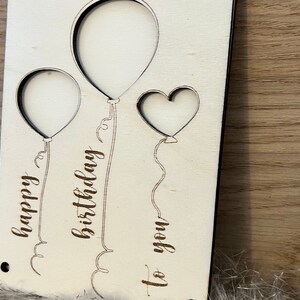 May include: A wooden birthday card featuring two balloon shapes and a heart cut-out. The words "happy birthday to you" are etched into the wood. The card is light-coloured with a natural wood grain.