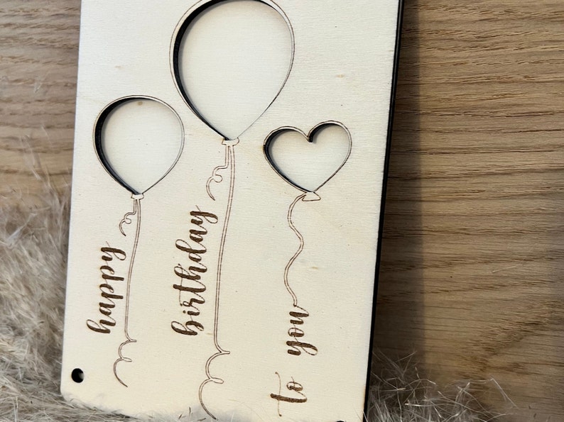 May include: A wooden birthday card featuring two balloon shapes and a heart cut-out. The words "happy birthday to you" are etched into the wood. The card is light-coloured with a natural wood grain.