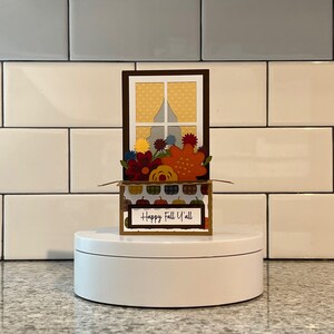 May include: A decorative fall-themed card featuring a window design with colorful paper flowers and pumpkins. The card includes the text "Happy Fall Y'all" and is displayed on a white platform.