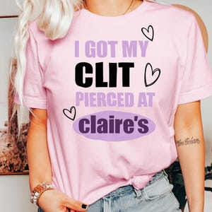 May include: A light pink t-shirt with the text "I GOT MY CLIT PIERCED AT claire's" in black and purple. The shirt has three heart graphics. The shirt is a short-sleeved crew neck.