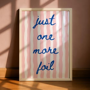 Peut inclure: Une affiche encadrée avec un fond à rayures roses et blanches. Les mots "just one more foil" sont écrits en bleu en écriture cursive. Le cadre est de couleur beige clair, et l'affiche est appuyée contre un mur.