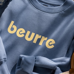 May include: A blue sweatshirt with the word "beurre" in yellow block letters. The sweatshirt has a crew neck and long sleeves. The fabric appears to be a soft, comfortable material.