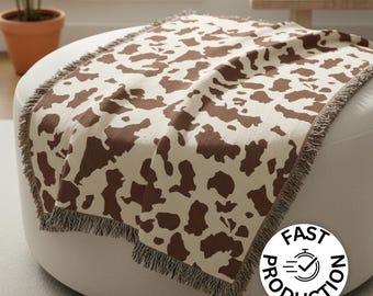 Op de afbeelding: Een crèmekleurige sprei met een bruin koeienprintpatroon. De deken heeft een gefranjerde rand en is over een witte poef gedrapeerd. De afbeelding bevat ook een plant in een pot en een kleine tafel.