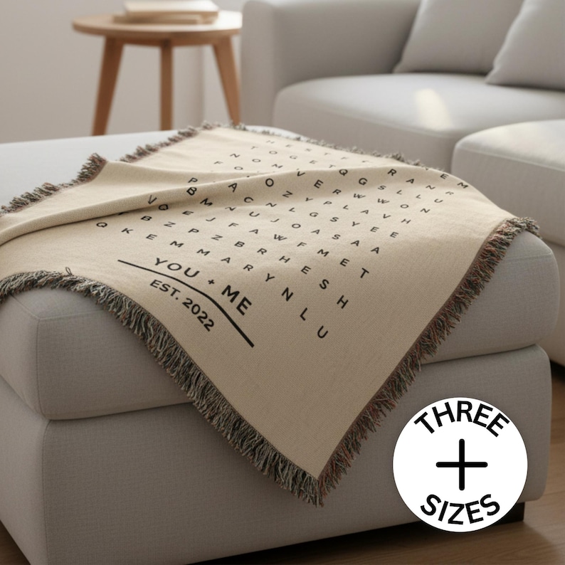 May include: A beige throw blanket with a word search design and fringed edges. The blanket features the text "YOU + ME EST. 2022" in black. A white circle with the text "THREE + SIZES" is in the bottom right corner.