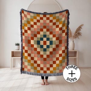 May include: A woven throw blanket featuring a geometric checkerboard pattern in shades of terracotta, mustard yellow, sage green, and cream, with some squares displaying a floral print. The blanket has fringed edges and text reading THREE SIZES.