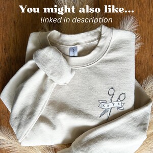 Puede incluir: Sudadera de color crema con el texto "You might also like... linked in description" en la parte superior. La sudadera presenta un gr&aacute;fico de tijeras con el nombre "KATE" en una pancarta. La sudadera est&aacute; doblada sobre una superficie de madera.