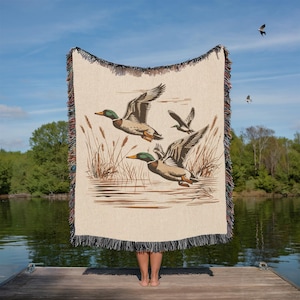 May include: A beige woven throw blanket featuring a detailed illustration of three ducks in flight over water and reeds. The blanket has a black fringe border with colorful accents. The background shows a lake and trees.