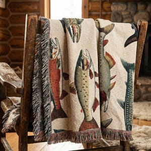 May include: A beige throw blanket draped over a wooden chair, featuring a repeating pattern of colorful fish illustrations. The blanket has a fringed edge with brown, red, and green accents, creating a rustic, outdoorsy aesthetic.