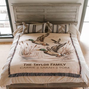 May include: A beige throw blanket with a duck illustration and the text "THE TAYLOR FAMILY" with names below. The blanket has a fringed edge and is displayed on a bed with striped pillows and a wooden headboard.