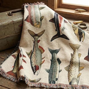 Puede incluir: Una manta beige con un patrón repetido de ilustraciones de peces en tonos rojos, negros y marrones. La manta tiene un borde con flecos y está sobre una superficie de madera. Se ve una caja de aparejos de metal vintage.