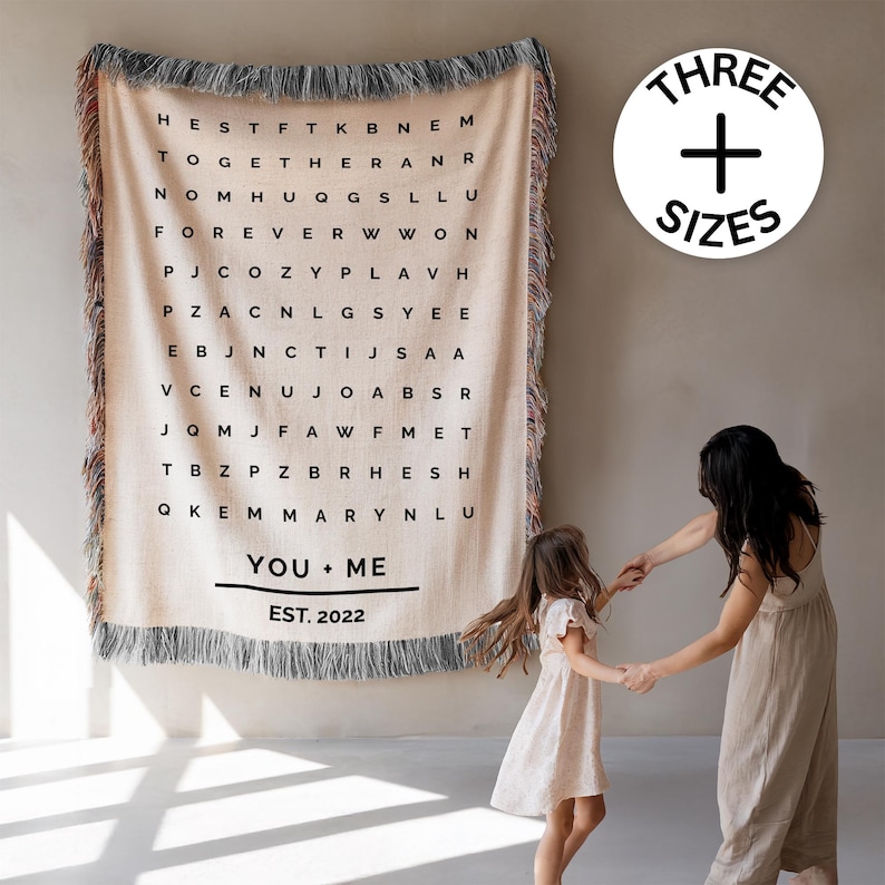 May include: A beige woven throw blanket with a word search design. The blanket has a fringe border and the words "YOU + ME" and "EST. 2022" are printed at the bottom. A circular graphic with the words "THREE + SIZES" is in the upper right.
