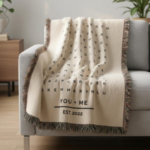 May include: Cream-colored throw blanket with a black letter design and fringe trim. The blanket features the text "YOU + ME" and "EST. 2022". The blanket is draped over a gray sofa.